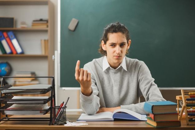 thinking young male teacher points at up sitting at desk with school tools on in classroom