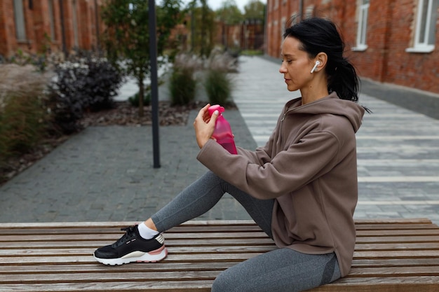 Thirsty fitness woman in earphones sitting on the bench and opening bottle