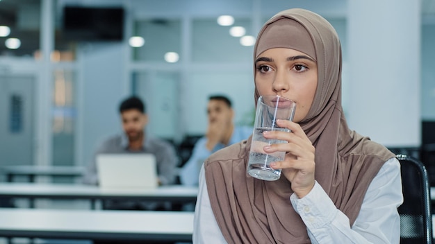 Thirsty millennial indian arabic female worker in hijab holding glass drinking pure mineral water in office thoughtful dehydrated woman employee enjoying fresh aqua Concept of healthy lifestyle