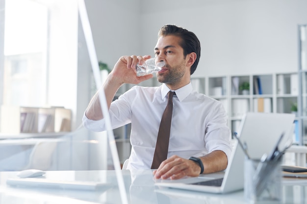 Thirsty young broker having glass of water while sitting in front of computer monitor in office