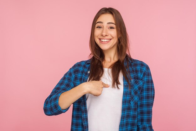 This is me. Portrait of charming happy girl in checkered shirt smiling and pointing at herself, being proud of own success, boasting of achievement. indoor studio shot isolated on pink background