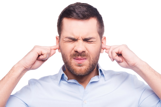 This is too loud! Frustrated mature man in shirt holding fingers in his ears and keeping eyes closed while standing against white background