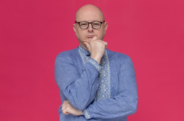 Thoughtful adult slavic man in blue shirt wearing optical glasses holding his chin and 