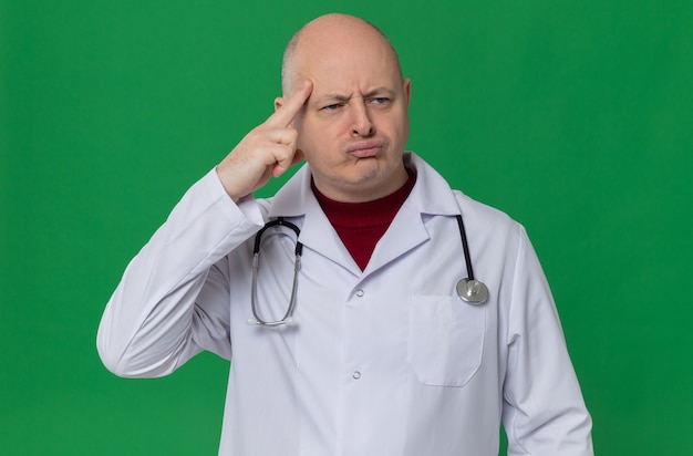 Thoughtful adult slavic man in doctor uniform with stethoscope putting finger on his temple and looking at side