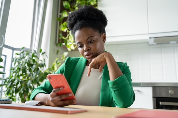 Thoughtful African American woman freelancer with phone in hand sits at work desk in home office