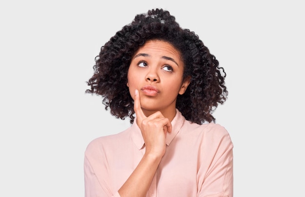 Thoughtful African American woman has puzzled expression frowns her face and looking at one side with hand on chin Dark skinned female cant make choice has questioned expression isolated on white