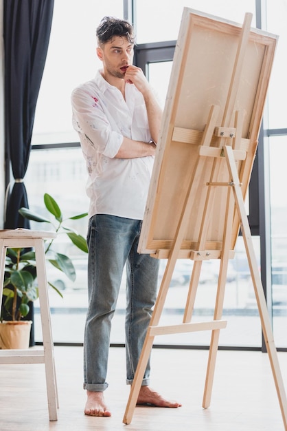 Thoughtful artist in white shirt and blue jeans standing at easel in gallery