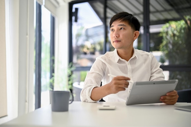 Thoughtful Asian businessman daydreaming about his career success while using tablet