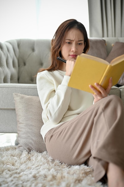 Thoughtful Asian female focused reading a book or adventure fiction book in her living room