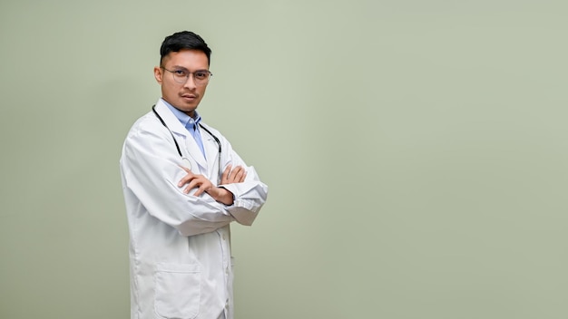 Thoughtful Asian male doctor in a white gown stands with arms crossed against an isolated background