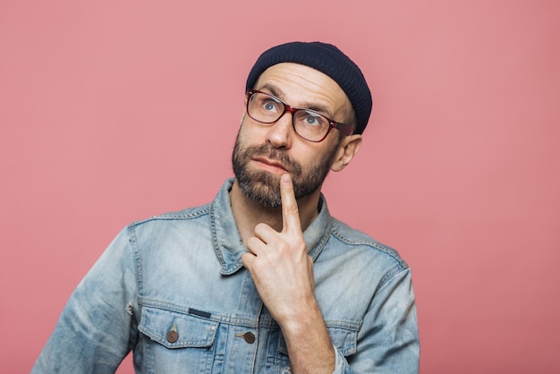 Thoughtful bearded male being deep in thoughts looks with pensive expression into camera concentrated on something poses against pink background People facial expressions daydreaming concept