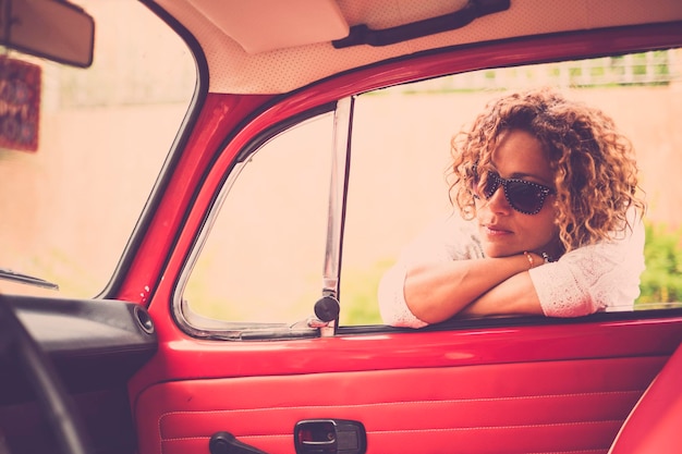 Thoughtful and beautiful caucasian middle age curly blonde woman smile and enjoy the outdoor leisure activity near her red vintage classic car ready to travel and live the day