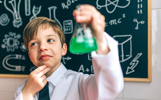 Thoughtful boy dressed as chemist with flask in front of a blackboard with drawings