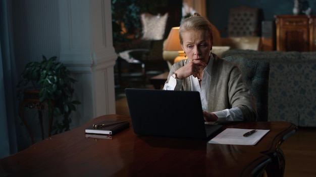 Thoughtful business woman working computer in vintage cabinet Senior pensive woman typing laptop computer in luxury home Focused old lady looking laptop in classic interior