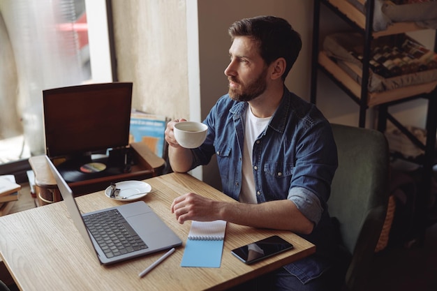 Thoughtful caucasian handsome man drinking coffee in cafe and working at laptop coffee break