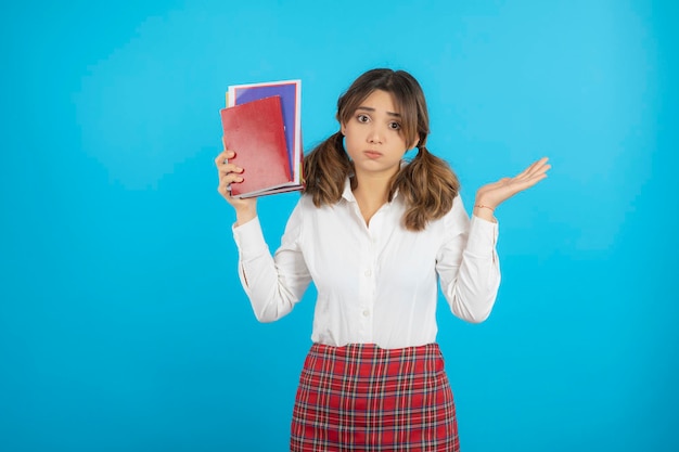 Thoughtful college girl holding her books up and stand on blue background. High quality photo