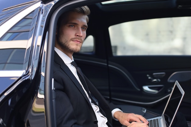 Thoughtful confident businessman sitting in the lux car and using his laptop.