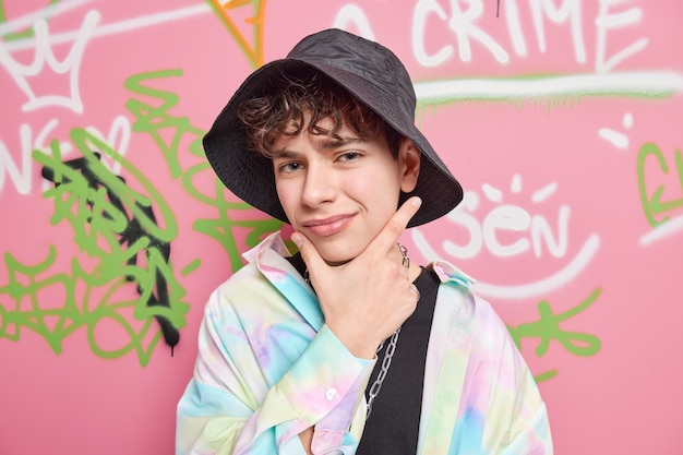 Thoughtful cool hipster guy holds chin considers something has displeased expression wears hat and shirt poses against graffiti wallmakes assumptions