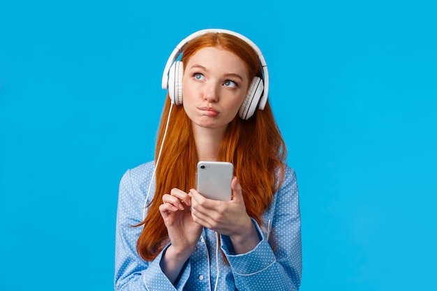 Thoughtful and creative, pensive caucasian redhead woman, foxy girl in nightwear, looking up thinking, wearing headphones listen music or podcast, holding phone, blue wall