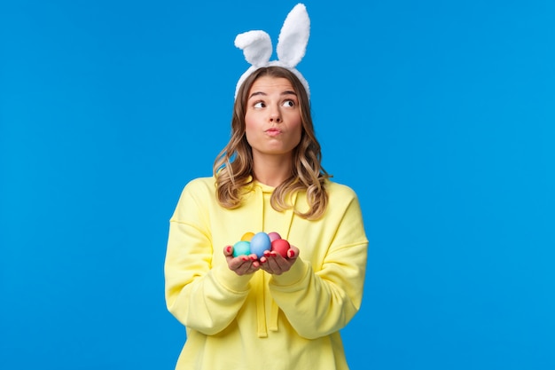 Thoughtful cute caucasian girl in rabbit ears thinking, looking up pondering and dreamy, holding Easter eggs, decide where hide it during traditional holiday game, on a blue wall