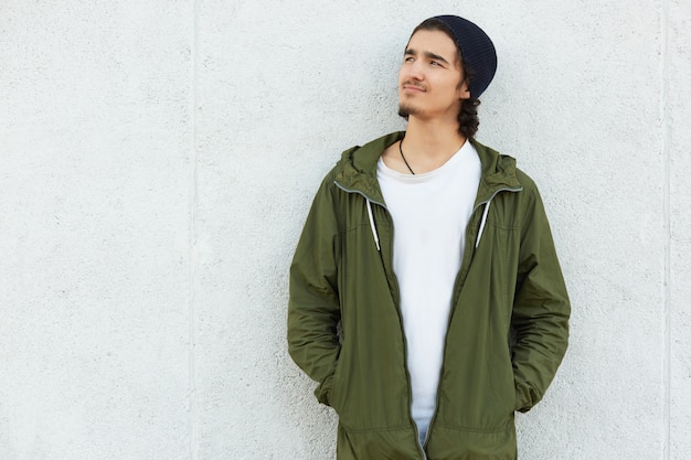 Photo thoughtful dreaming mysterious young man with curly hair wearing white t shirt, black trendy hat and green jacket