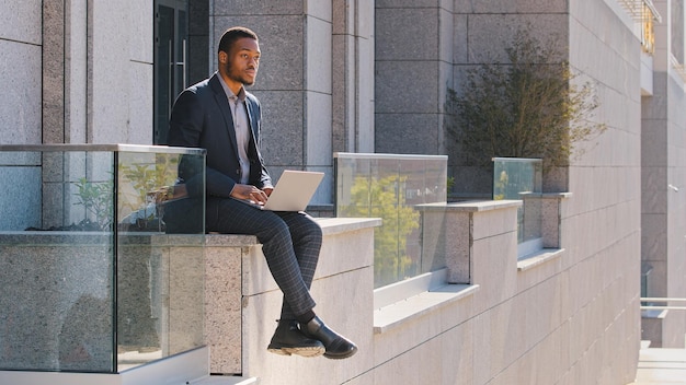 Thoughtful ethnic businessman African American man manager freelancer sitting outdoors typing on laptop concerned about problem worker guy thinking difficult decision looking away planning strategy