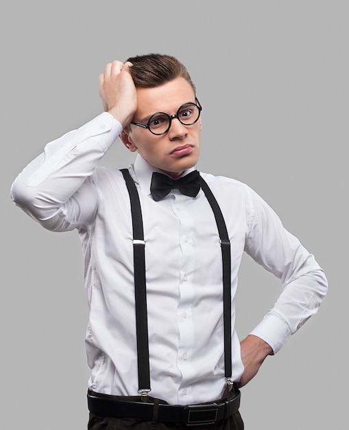 Thoughtful geek. Portrait of thoughtful young man in bow tie and suspenders holding hand in hair and looking away while standing against grey background