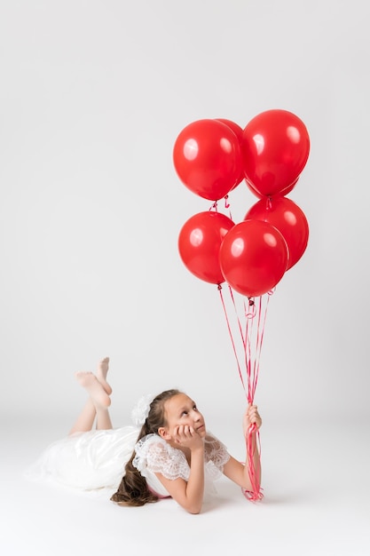 Thoughtful girl ten years old dressed in white dress holding lot of red balloons in hand looking up