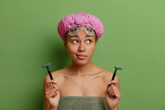 Thoughtful healthy young Afro American woman female model wears bath hat wrapped in soft towel holds two razors for shaving isolated over vivid green wall