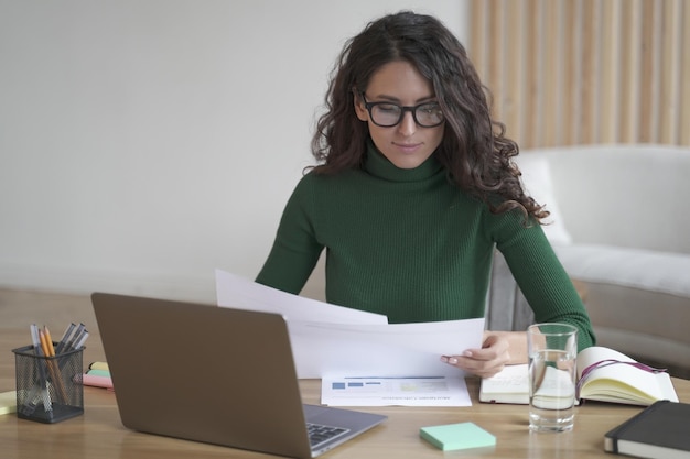 Thoughtful italian female freelancer in glasses sitting at desk holding papers and preparing report