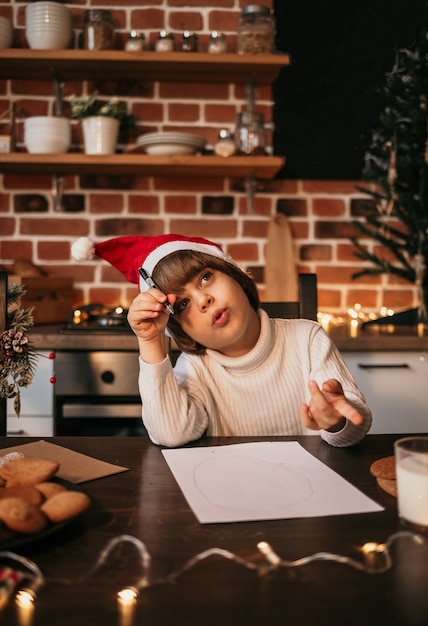 A thoughtful little boy in a white knitted sweater and a red hat is sitting at a table and writing a New Year's letter