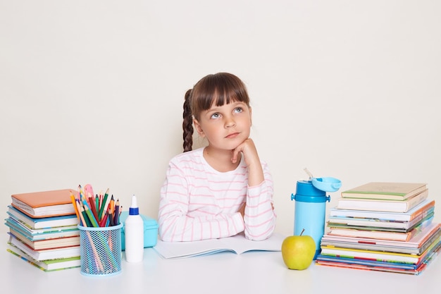 Thoughtful little schoolgirl wearing striped shirt sitting at the desk surrounded with books and other school supplies looking away thinking during lesson or while dong homework holding chin