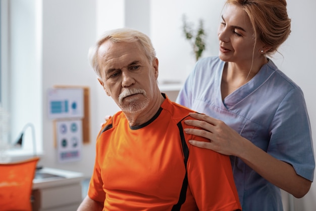 Thoughtful look. Pleasant aged man sitting in front of his nurse while thinking about his health