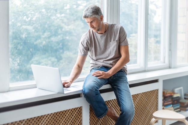 Thoughtful man with a laptop sitting on the windowsill