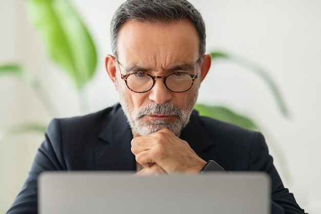 A thoughtful mature man in a smart casual suit with glasses deeply concentrating on his laptop