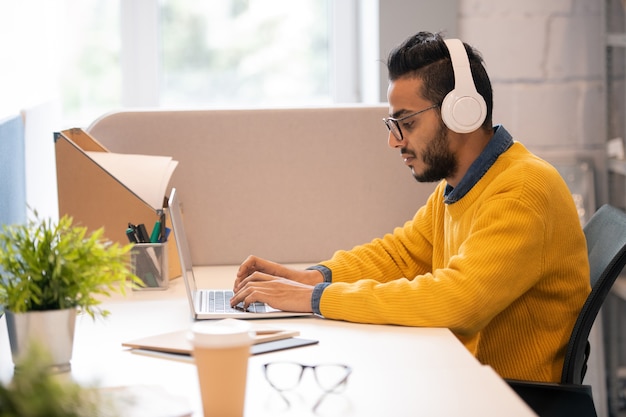 Thoughtful modern young Arabian marketer with beard sitting at desk and listening to music in wireless headphones while working with laptop