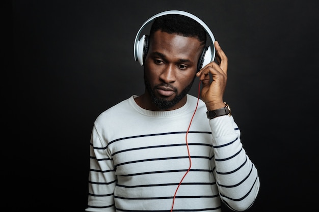 Thoughtful nice man enjoying music while using headphones and standing isolated in black wall