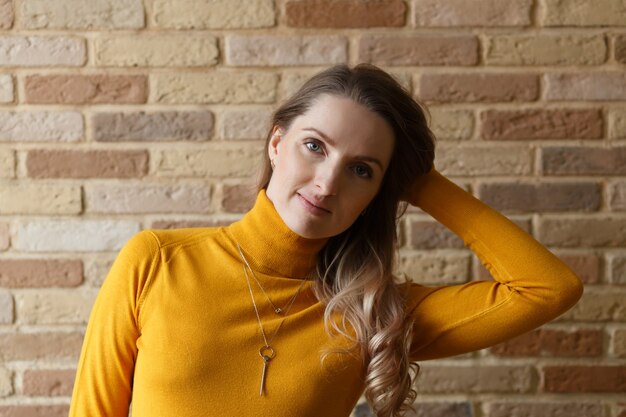 Thoughtful pregnant woman in yellow cardigan caressing her belly and posing near by brick wall