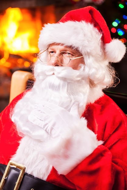 Thoughtful Santa. Thoughtful Santa Claus sitting at his chair and touching beard with fireplace and Christmas Tree in the background