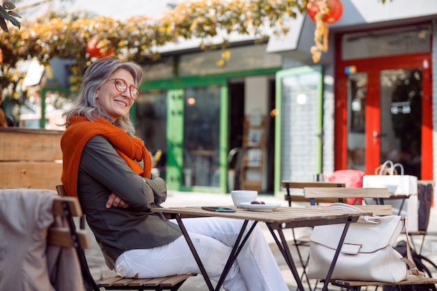 Thoughtful senior lady with crossed arms sits at table on outdoors cafe terrace