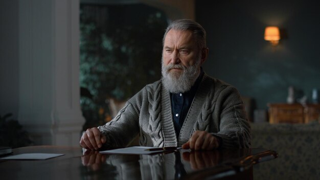 Thoughtful senior man sitting with crossed arms at table with papers indoors Serious businessman thinking in classic interior Elderly man working with documents at luxury home