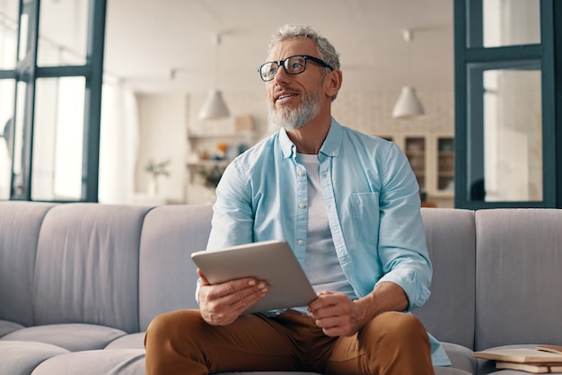 Thoughtful senior man using digital tablet and looking away with smile while sitting on the sofa at home