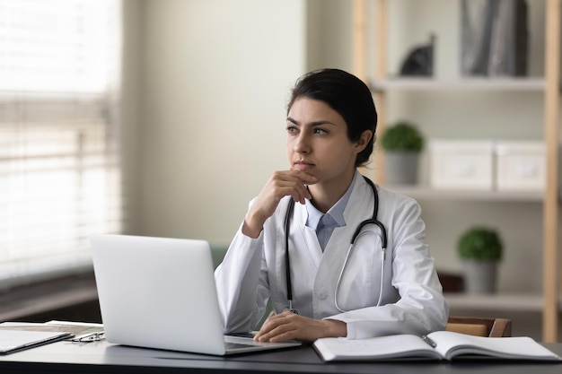 Thoughtful serious Indian female doctor physician touching chin sitting at work desk in office with laptop looking in distance pensive practitioner in white uniform with stethoscope thinking