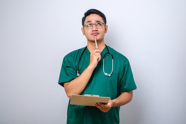 Thoughtful smart asian nurse in scrubs looking away and thinking while holding clipboard and putting pen on his chin