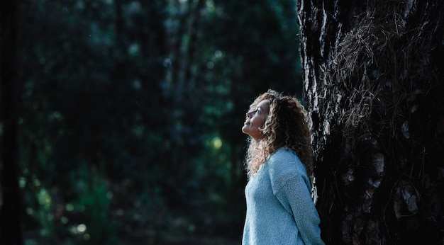 Thoughtful woman standing against a trunk tree in the forest looking up Concept of environment and dark woods with people Global warming and nature park protection Tourist in the woods outdoor