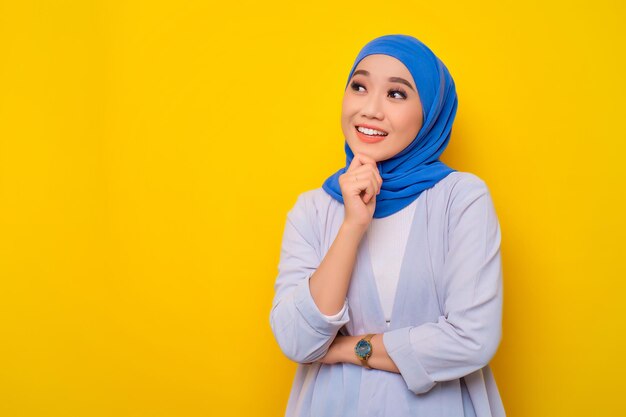 Thoughtful young Asian Muslim woman looking up at copy space having new idea isolated on yellow background