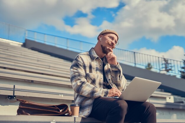 Thoughtful young bearded man with mustache works outisde while holds laptop on legs city space