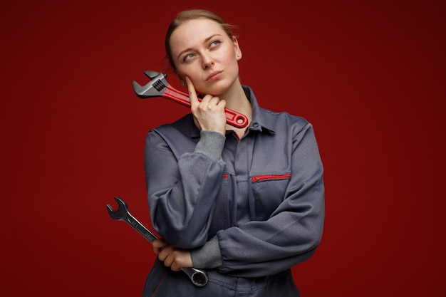 Thoughtful young blonde female engineer wearing uniform holding wrenches keeping finger on cheek looking up 