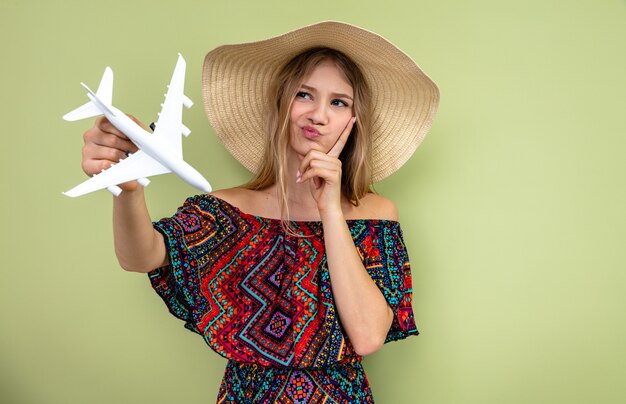 Thoughtful young blonde slavic woman with sun hat holding plane model and looking at side 