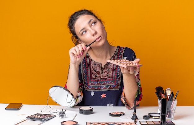 Thoughtful young brunette girl sitting at table with makeup tools holding eyeshadow palette and makeup brush looking at side isolated on orange wall with copy space